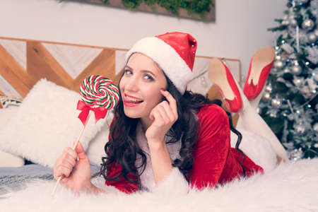 Sexy santa woman licking lollipop on the background of the Christmas tree. A sweet Christmas present. Pretty woman has fun on the bed.の写真素材