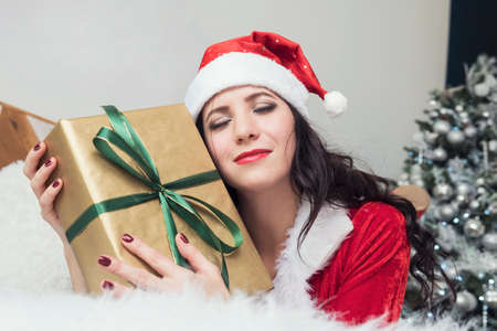 Smiling teenage girl in santa helper hat with gift box on the background of the Christmas tree at home in the morning light.. Positive emotional Santa girl. Xmas. Girl hugging gifts.の写真素材