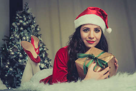 Cute cute girl in clothes santaclaus holding a gift with a green ribbon lying on the bed against the Christmas tree. New year and Christmas celebrationの写真素材