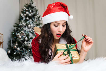 Excited young woman opening a Christmas gift. Cute girl in a Santa Claus costume unleashes a ribbon on a new year gift lying on the bed on the background of a decorated Christmas treeの写真素材