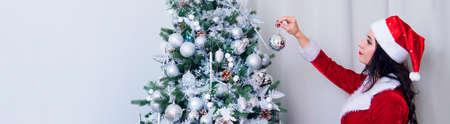Beautiful woman in a sexy Santa Claus costume decorate the Christmas tree decorations at home. Girl hangs a silver ball on a green Christmas tree. Preparations for the new year celebrationの写真素材