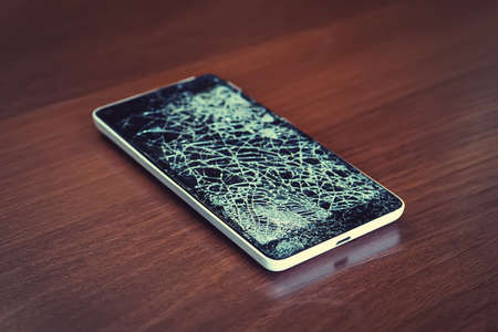 Black smartphone with broken screen on the wooden table. Phone repair concept. Old broken phone with black screen. The display cracked in the form of a web.の写真素材