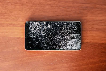Modern frameless Smartphone with broken screen on the wooden table. Phone repair concept. Old broken phone with black screen. The display cracked in the form of a web.の写真素材
