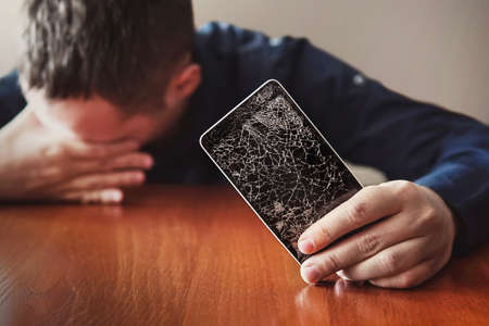 Guy with bristles, is showing his hand on a smartphone. Broken screen of modern frameless phone. guy crying with his eyes closed and hands in his face.の写真素材