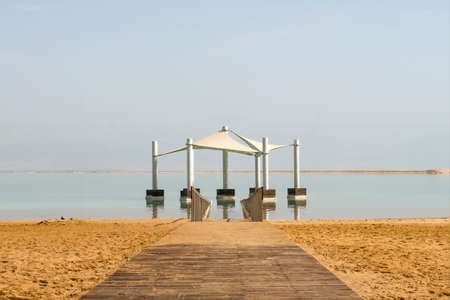 Wooden path on yellow sand leaving in the distance of the dead sea. The path under the canopy in salt water. Canopy from the sun in the water for swimming in dead sea. Ein Bokek Israel.の写真素材