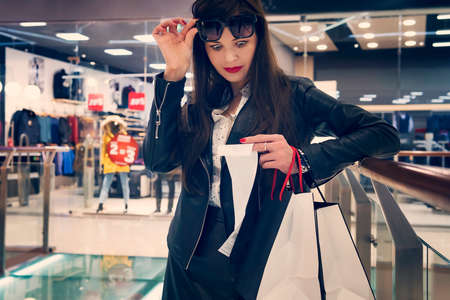 Portrait of young woman with big eyes staring in amazing at her bill at shopping center background. Lady taking off her dark glasses to have a good look at total price in bill. Girl spent too muchの写真素材