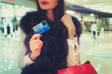 Half-face shot of young chic woman with red lips in shopping centre holding credit card and bag in hands. Trendy brunette girl is going to do shopping using payment card. Lady in fur jacket in storeの写真素材
