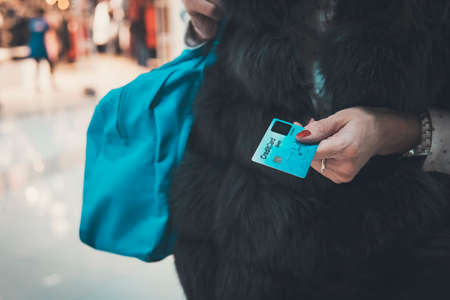 Well-dressed lady in fur holding credit card and bag at blurred mall background. Woman paying in fashion boutique giving bank card to cashier. Female shopping time. Desire to spend money and consumeの写真素材