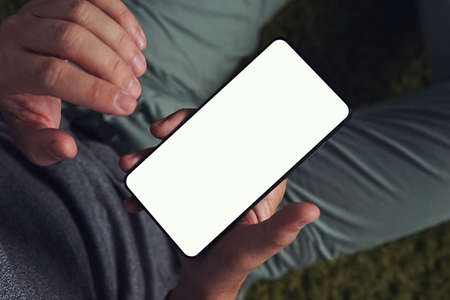 A slantwise top-down view of a young man using the smartphone with the white screen. Man holds the smartphone in his right hand and ready to touch the screen with the left hand. Person is on-line.の写真素材