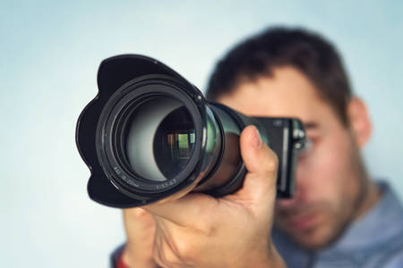 male photographer hold camera in his hands. Mirrorless camera close up in the hand of a young man on blue background. The reporter's taking a picture. the photographer looks into the camera. close-upの写真素材