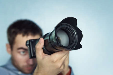 Close up photo of man in hat on blue background taking a photo with digital mirrorless camera. Young cute male photo reporter with a small camera in her hands. Paparazzi taking a picture.の写真素材