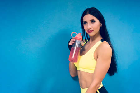 Close up of athletic girl in yellow sport top standing at blue wall and holding shaker with energy drink. Weightloss drinking after workout in gym. Attractive brunette woman holding protein drinkの写真素材