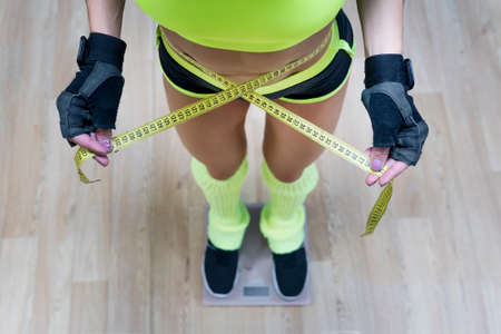 Top view of pretty slim woman with measure around her body standing on scales. Fitness woman dressed in sport outfit measuring her weight and waist size. Athletic girl looks at results of her workoutの写真素材