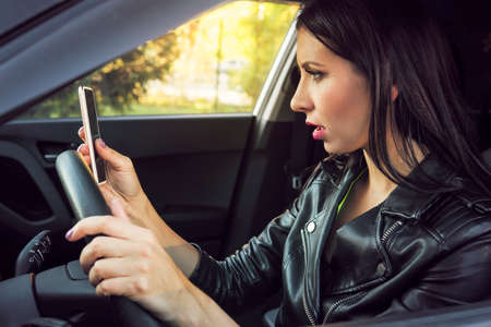 Brunette woman sitting inside car with mobile phone in hand texting while driving. Shocked girl checking her smartphone not paying attention at road stunned by bad text message. Dangerous distractionの写真素材