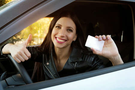 Young girl with beautiful smile sitting in white car showing an empty white card and thumbs up hand sign. Woman has got driving license and is very happy. Pretty girl at wheel inside car is excitedの写真素材