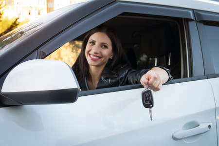 Happy smiling businesswoman showing keys of her new expensive vehicle. Front window view to satisfied female driver holding and demonstrating car keys. Lady is lucky to purchase a car. New ownerの写真素材