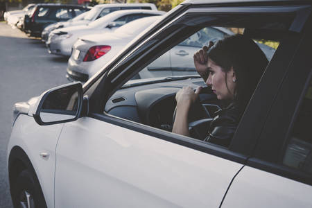 Side view of beautiful young woman looking in car side-view mirror sitting in vehicle. Female driver preens herself looking in car mirror. Girl while away the time waiting in white automobileの写真素材