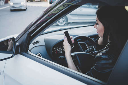 Woman using navigation app on smartphone while driving a car. Side view of brunette girl sitting in vehicle and looking at blank screen of cellphone. Female driver using device at wheel. Girl drivesの写真素材