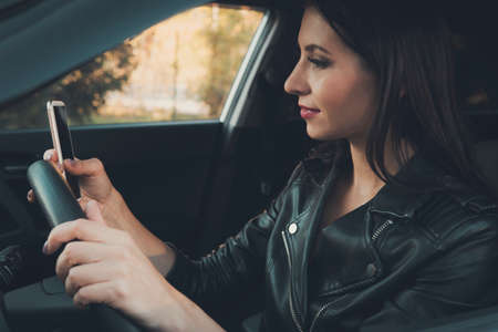 Smiling young woman using her mobile phone while driving. Attractive girl sitting inside a car and looking at her mobile black screen. Female checking her cellphone while waiting in a vehicle. Gadgetの写真素材