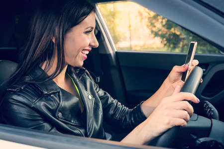 Happy smiling woman holding her smart phone sitting inside a car. Female driver laughing reading message and chatting in mobile. Girl looks at her cellphone screen and smiles while being at wheelの写真素材