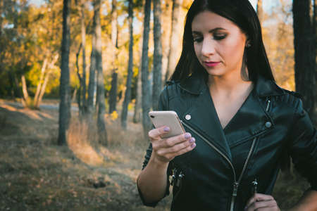Autumn walk. Technologies. Pretty girl in leather clothes is using smartphone while walking in the park. Girl stays online being in autumn park. Woman using cellphone at blurred nature backgroundの写真素材