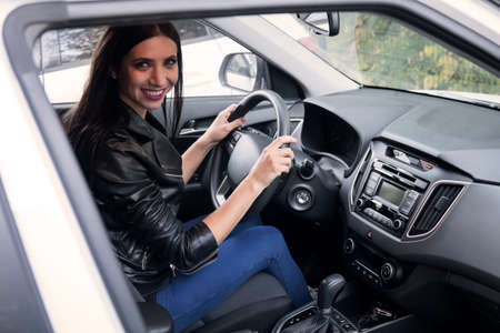 Beautiful smiling female driver in black jacket sitting inside car and holding steering wheel. A look into the vehicle window to happy attractive brunette driving automobile. Excited female driverの写真素材