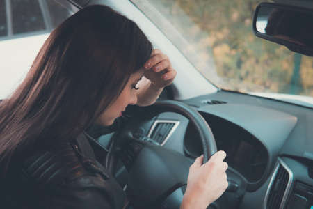 Closeup picture of young pretty lady waiting in car leaning on wheel with eyes closed on outdoors background. Girl feeling sleepy while waiting for somebody in her vehicle. Tired or depressed womanの写真素材