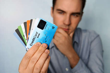 The young man is holding a stop of credit and debit cards in a pensive pose. The guy chooses a card to pay. businessman holds a large stack of credit cards in his hand and looks at themの写真素材