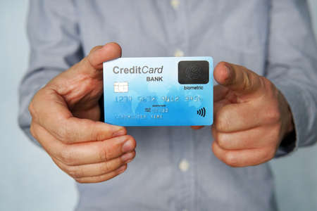 Young man stands at white background and holds credit card with fingerprint sensor in one hand and points with index finger of other hand to it. Purchasing with biometric technology. Safe method.の写真素材