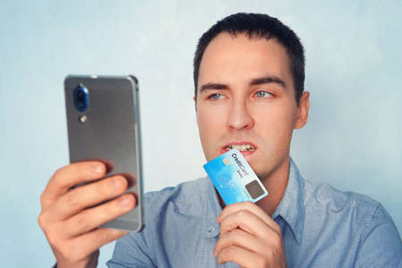 shot of male in shirt making online payment via plastic card and smart phone. man holding mobile and credit card in the teeth. Online banking. E-commerce. Purchase. man is nervous and afraid.の写真素材