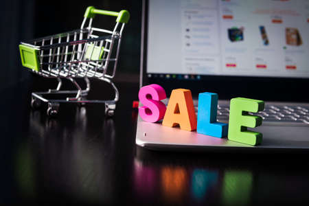 Many-colored wooden word SALE set on laptop bottom at the background of opened web page on the screen and small shopping trolley. Concept of shopping online. Good buy in internet. Sale in E-commerce.の写真素材