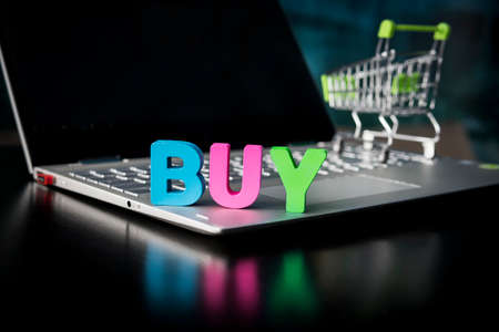 Creative shot of a wooden many-colored word BUY placed on laptop bottom at black screen and toy shopping trolley background. Concept of buying things in internet. Online shop promotion. E-business.の写真素材