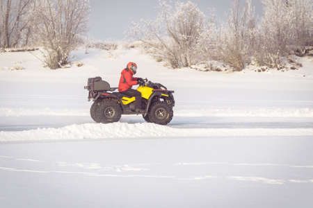 man driving a quad bike in the winter field. competition yellow ATV in the winter in the snow. The ATV rider in red uniform and helmet.の写真素材