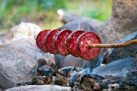 Roast smoked sausage over the fire. Sliced sausages. Sausage impaled on a stick. Fast dinner camping in the countryside. small campfire. Grilling sausages over an open fire outdoorsの写真素材