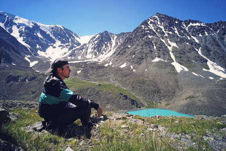 Young man in black sportswear is sitting on cliff's edge and looking to misty valley bellow. Beautiful background landscape with high mountains with snowy peaks.の写真素材
