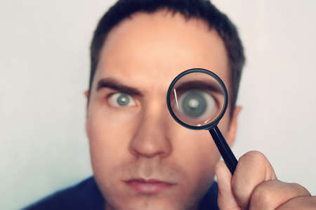 Man on white background curiously looks through the magnifying glass. Male fuzzy eye magnified through the loule. Person uses magnifier to look closely at some object. Detective search for evidenceの写真素材