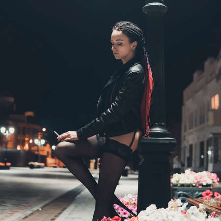 Sexy model stands under the lantern and poses in the image of a corrupt girl. Prostitute on the street smoking a cigarette with his knee bent. Girl addict in a leather jacket.の写真素材