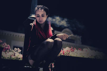 Young sexy girl sitting under a lantern on the street in the night city and Smoking a cigarette....
