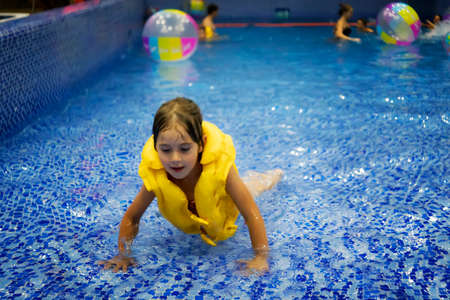 a little joyful girl in a yellow life jacket swims in the water in the pool in a closed water Park. The child learns to swim.の写真素材