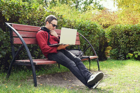 Young stylish male student texting message on laptop in park on autumn. The guy works at a laptop in the open air. Remote work. Thoughtful young man looking at the laptop screen.の写真素材