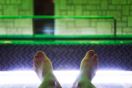 A close up of barefooted man lying down near indoor luxury swimming pool. A shot of young man's feet at the indoor pool background. Man feeling comfortable and relaxed after a swim in a spa pool.の写真素材