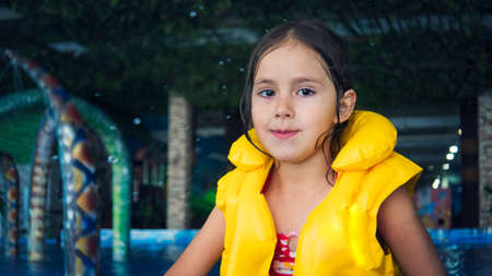 A close up of a cute smiling girl looking at camera at the water park background. A little smiley beautiful girl splashes water in indoor swimming pool. Water drops fall on the girl at aquapark pool.の写真素材