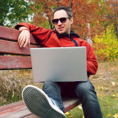 Joyful guy communicates through the Internet on the street. Programmer enjoy with notebook sitting in autumn park. Handsome man working on his laptop. Free work outside, freelance out of doors.の写真素材