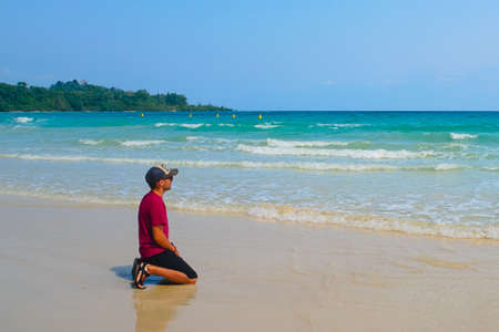 A young guy in a cap kneels on the sand in front of the turquoise sea. Exotic tropical island in the Indian ocean. Lost male tourist sitting on a beach and thinking.の写真素材