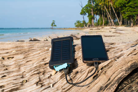 Battery solar energy device on a background of the sandy beach of an uninhabited island. Charge smart phone from the solar battery. the solar battery lies on a wooden logの写真素材