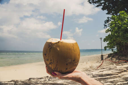 Female hand with pink nails holding a fresh coconut with coconut milk on the background of the beach and the sea and a cloudy sky on a tropical exotic island.の写真素材