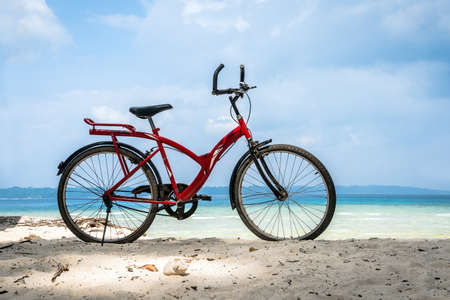 Red vintage bicycle on white sand beach over blue sea and clear blue sky background, spring or summer holiday vacation concept. Bicycle parked by the seaの写真素材