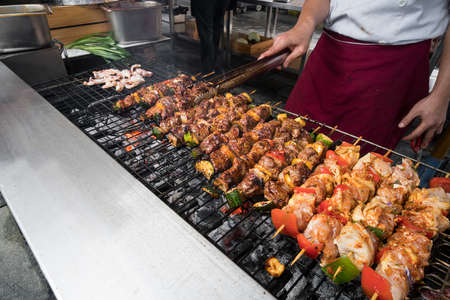 Food concept. Young handsome chef in white uniform monitors the degree of roasting and turns meat with the forceps in interior of restaurant kitchen. Preparing traditional beef on barbecue oven.の写真素材