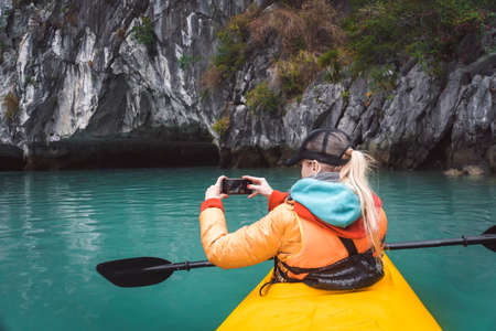 The girl takes a picture of a beautiful landscape sitting in a kayak.. kayaking tour of the islands, a holiday in Asia, a kayak's nose on the water. photograps in the boat.の写真素材