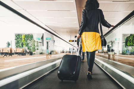Woman traveller with travel suitcase or luggage walking in airport terminal walkway for vacation travel abroad. concept of travel around world, tourism. Brunette in yellow skirt goes on escalator.の写真素材